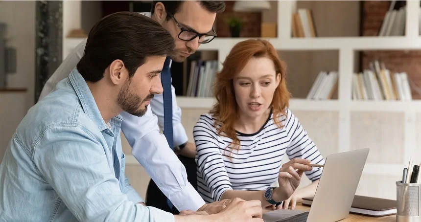 Three coworkers gathered around a laptop, discussing work in an office with a bookshelf background