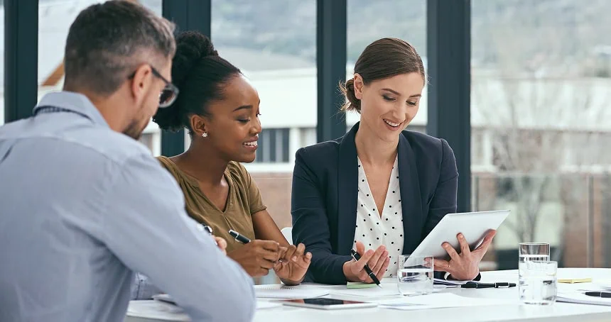 Three colleagues at a table reviewing a tablet during a meeting in a glass-walled office