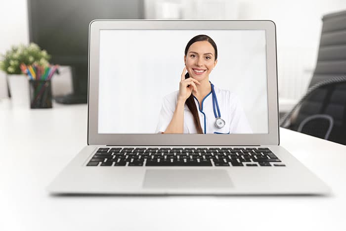 On a laptop during a videoconferencing session, the screen shows a smiling female doctor with a brown ponytail wearing white scrubs and a blue stethoscope around her neck touching her ear where headpiece connects to the audio.