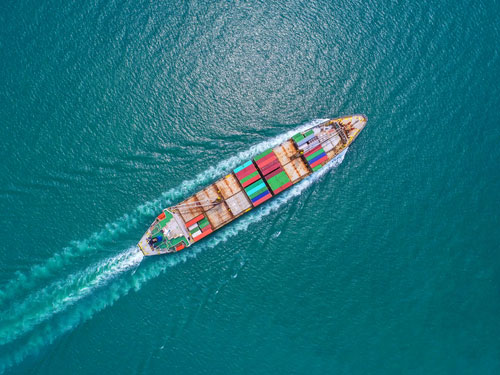 A boat shipping modern colorful cargo for transportation on the ocean headed diagonally up from bottom left to top right corner with a wake behind it