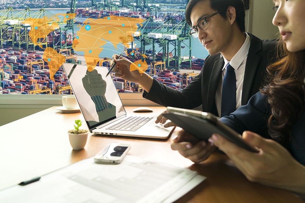 A woman and man sit in front of a laptop discussing transportation, shipping, and supply chain management. The image features a virtual overlay of a world map in orange. Outside the window is a dock with cargo ships and body of water in a city.